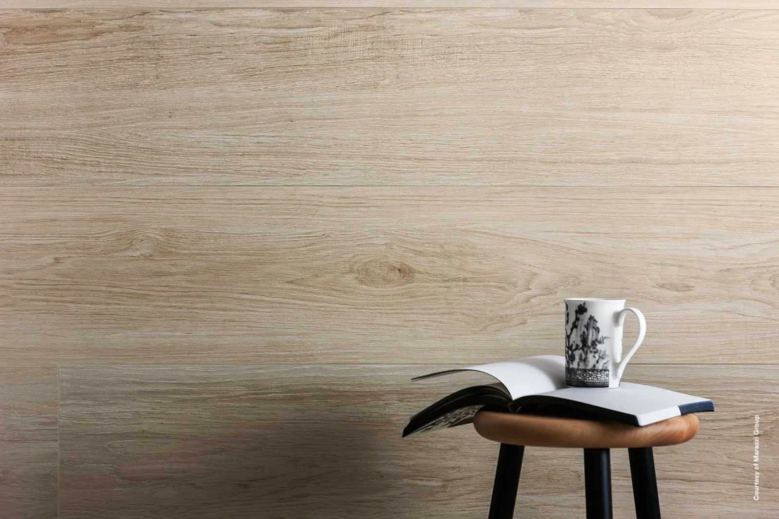 Nahansicht einer Wand aus hellen Holzoptik-Dielen, davor ein Holzhocker mit aufgeschlagenem Buch und bemalter Tasse.