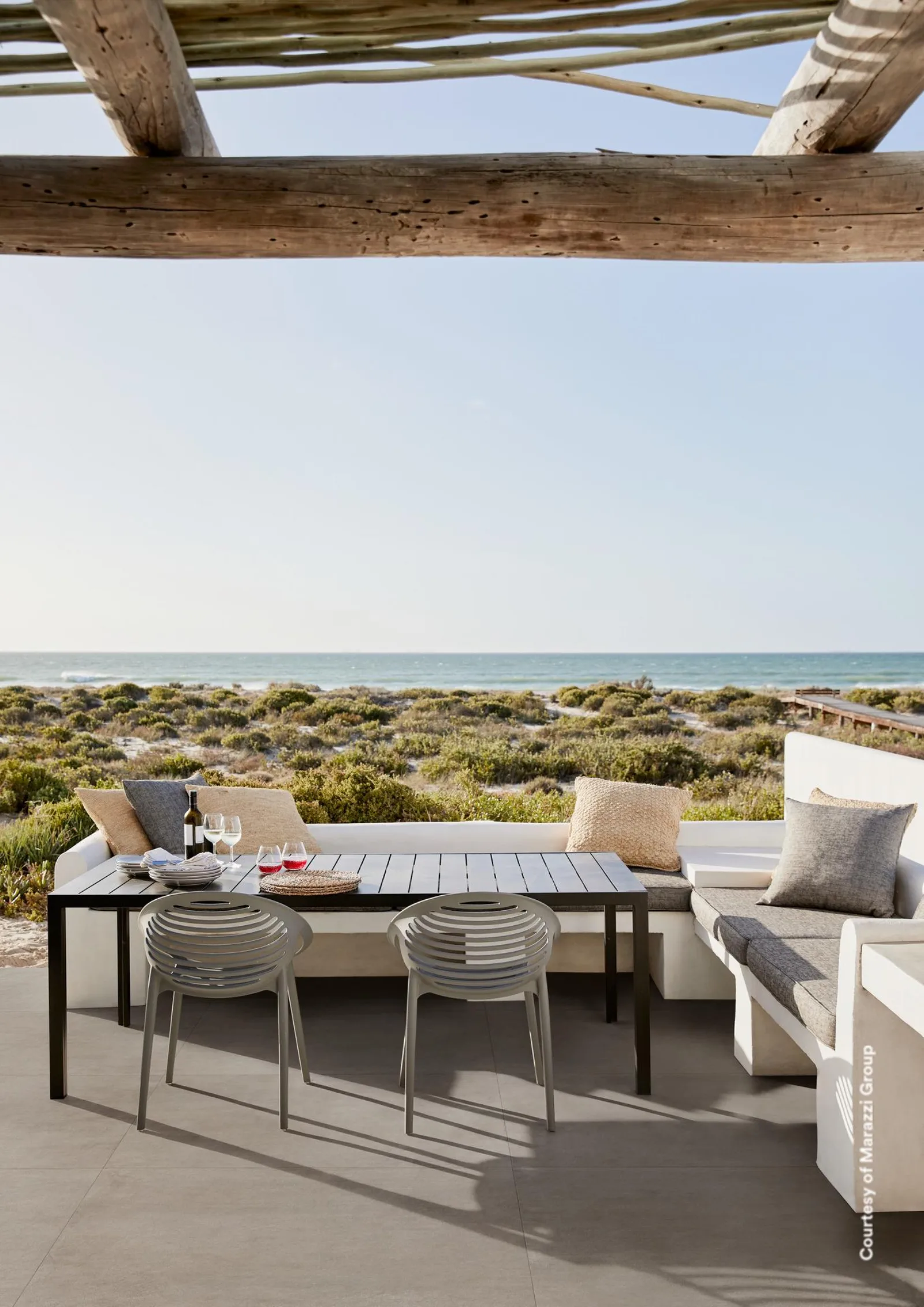 Überdachte Terrasse am Meer mit Esstisch und Sitzbank auf beigegrauem Betonoptik-Boden im Großformat.