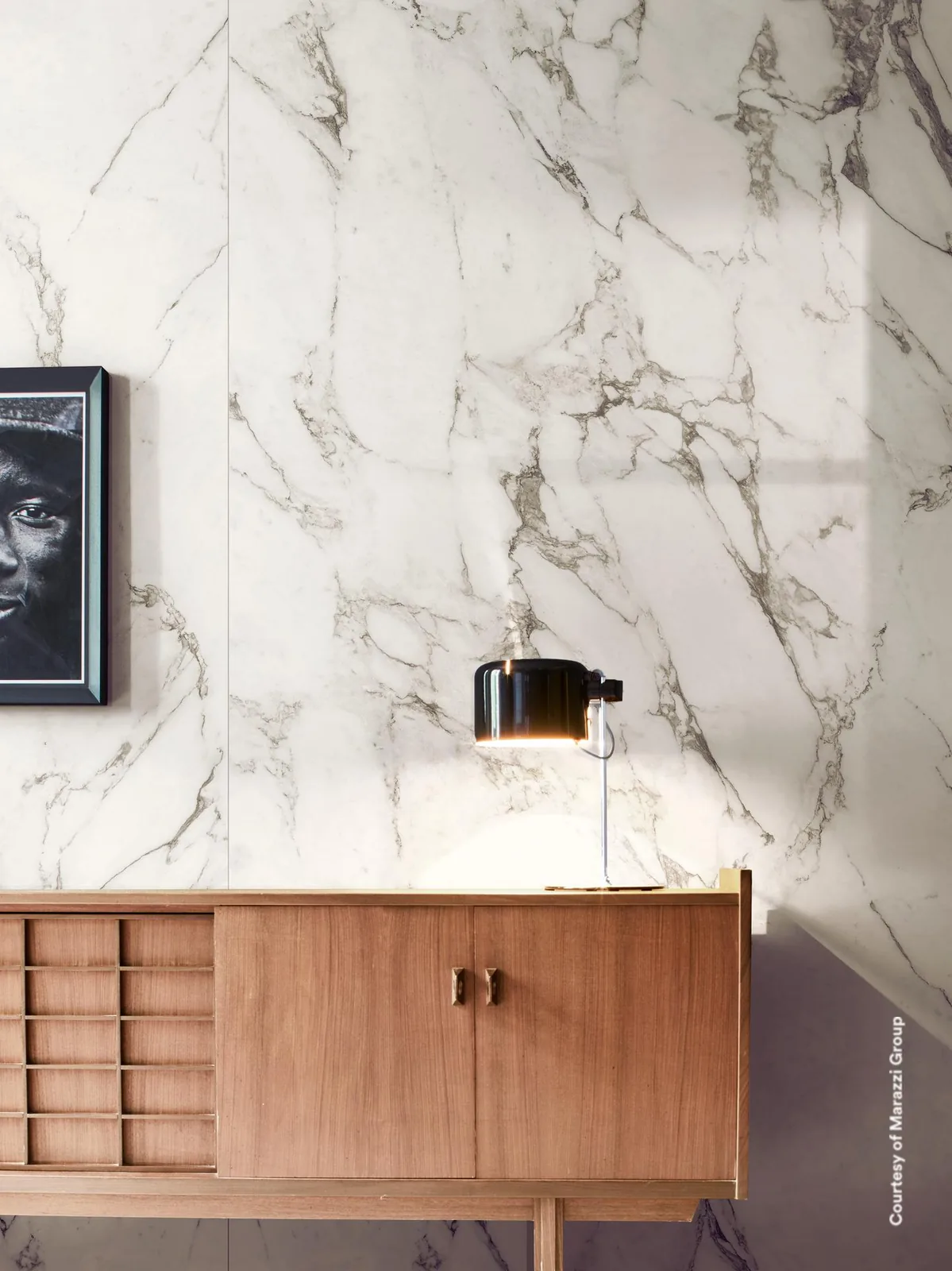 Wand in weißer Marmoroptik mit dichten grauen Adern hinter einem Sideboard aus Holz mit schwarzer Tischleuchte.