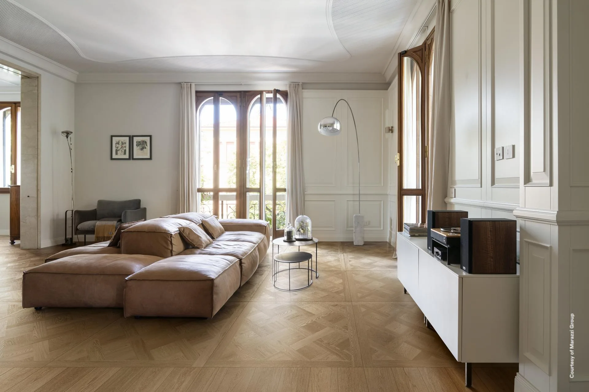 Wohnraum mit braunem Ledersofa und Bogenlampe vor hohen Bogenfenstern auf mittelbraunen Holzoptik-Dielen.