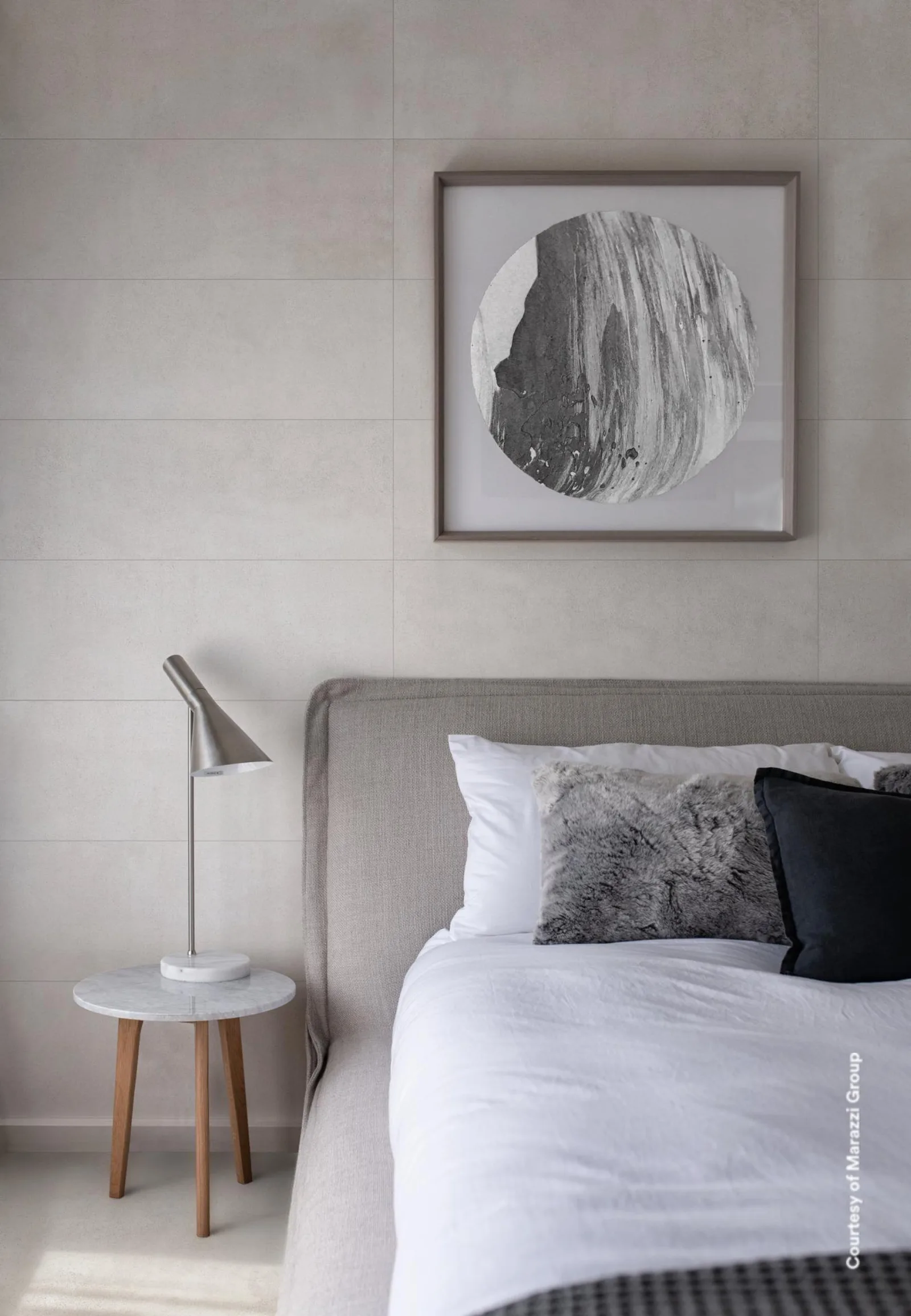 Bett mit grauem Kopfteil, kleiner Beistelltisch und gerahmtes Bild vor heller Wandfläche.