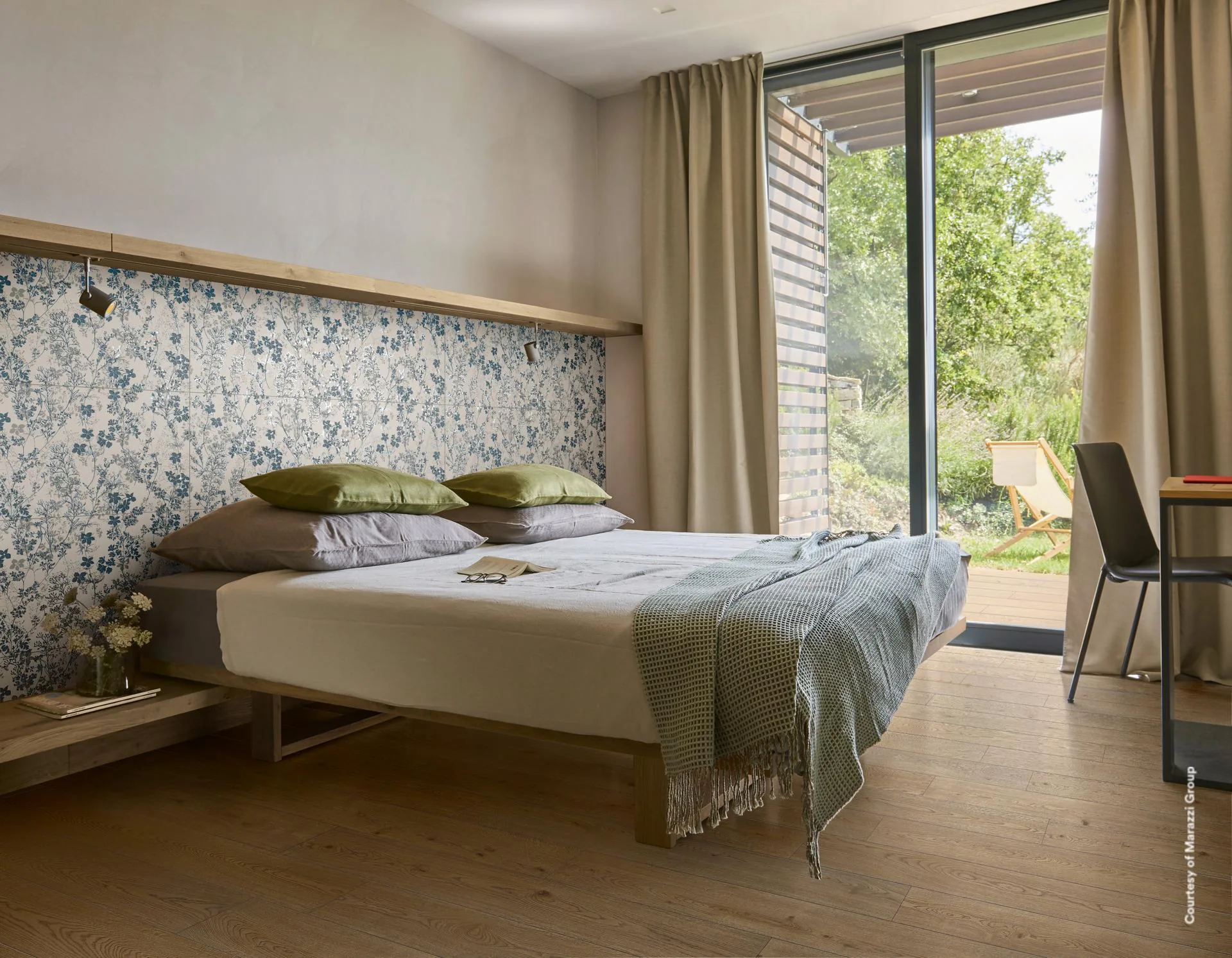 Schlafzimmer mit blau-weiß geblümter Wand hinter dem Bett, Holzboden, grünen Kissen und Blick durch Glasfront in den Garten